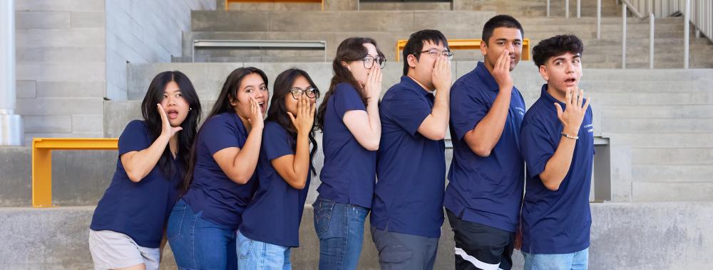 7 Program Assistants pose in a line, pretending to play "Telephone".
