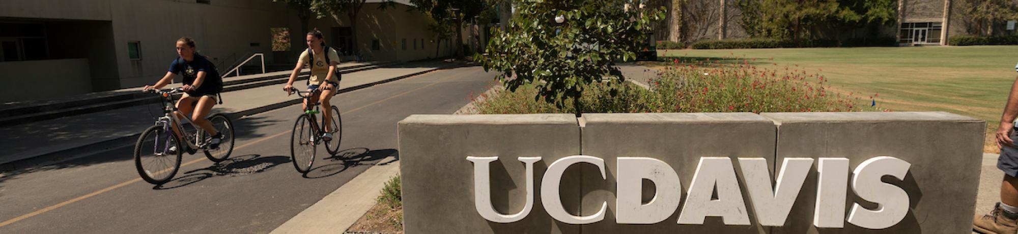 Two students bike by a UC Davis sign.